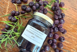 A glass jar of Blueberry Tarragon Jam from Timber House Resort is displayed on a rustic wooden table, surrounded by fresh blueberries and fresh tarragon sprigs.